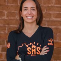 Sabine Zink Bolonhini é uma mulher branca de cabelos castanhos e sua uma blusa preta com letras laranja