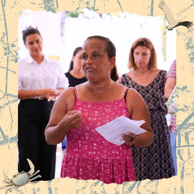 Ana Paula Santos é uma mulher negra não retinta, está de cabelo preso, usando um vestido rosa e em meio a outras mulheres