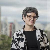 Carol é uma mulher branca de cabelos curtos grisalhos, ela usa óculos, uma blusa preta e um blazer branco com estampa preta