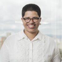 Daiene é uma mulher negra de pele clara e cabelos pretos curtos, ela usa óculos e uma camisa branca