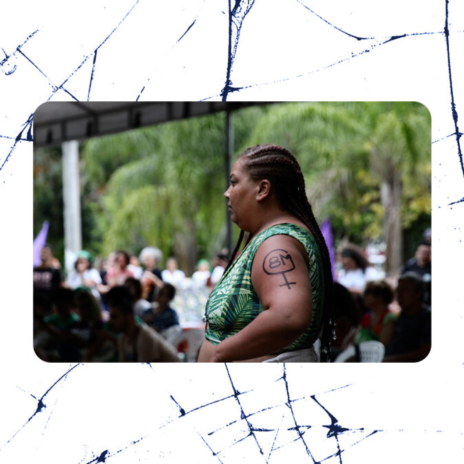 Mulher negra com tranças e roupa estampada em verde aparece de perfil com o símbolo feminino e 8M desenhado no braço, durante ato público com outras pessoas ao fundo.