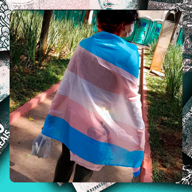 Samuel* está de costas usando máscara e com a bandeira do orgulho trans enrolada nos ombros, caminhando por uma calçada em área arborizada, com banheiros químicos ao fundo. A imagem representa resistência e afirmação de identidade
