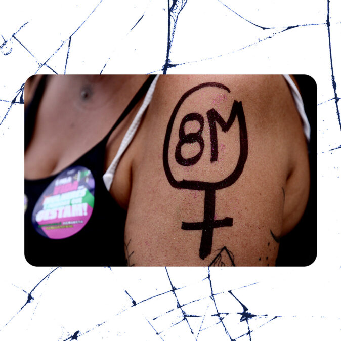 Braço de uma mulher com símbolo do feminino desenhado em tinta preta e a sigla "8M" dentro dele, em referência ao 8 de março, Dia Internacional das Mulheres. Ela usa um adesivo de participação em ato feminista.