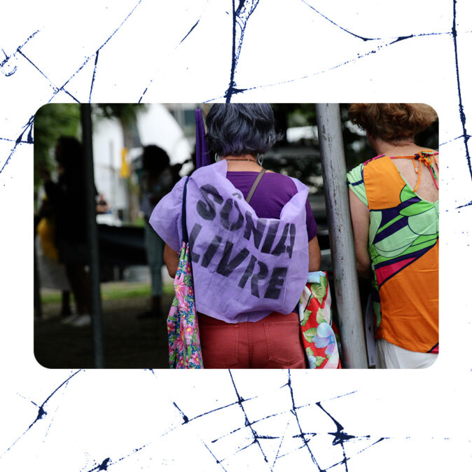 Mulher de cabelos grisalhos veste pano lilás com os dizeres “Sônia Livre” nas costas, durante ato público ao ar livre, ao lado de outra mulher com roupa colorida.