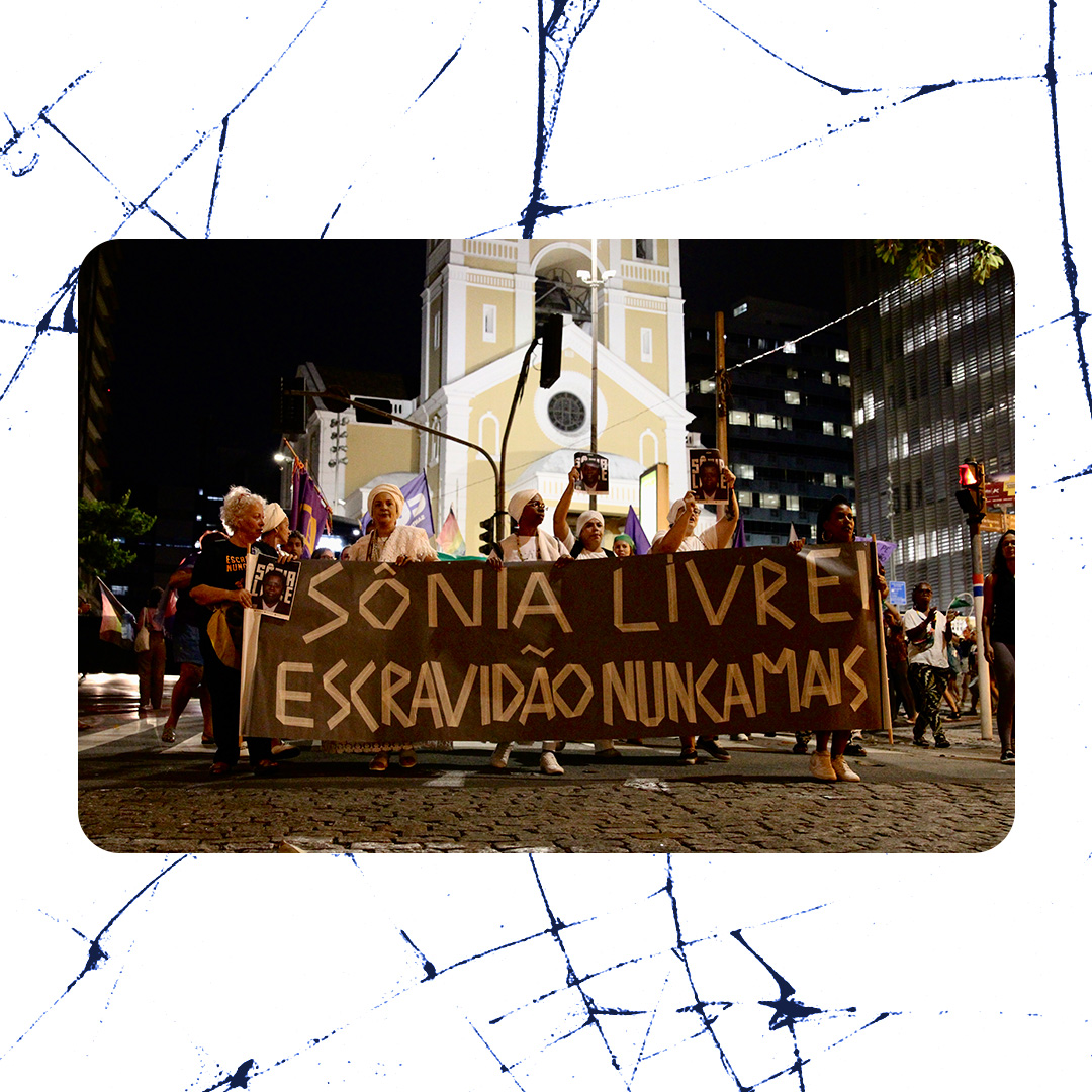 Manifestantes marcham à noite em uma avenida segurando faixa com os dizeres "Sônia Livre, Escravidão Nunca Mais", em frente a uma igreja iluminada, durante protesto por justiça para Sônia Maria de Jesus.