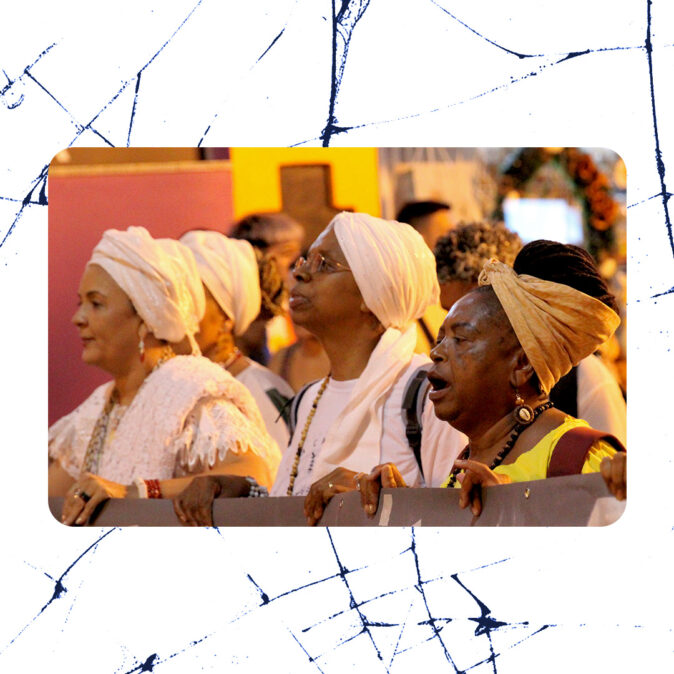 Mulheres negras vestidas de branco e amarelo, com turbantes e colares, participam de ato noturno segurando uma faixa, em manifestação pela liberdade de Sônia Maria de Jesus.