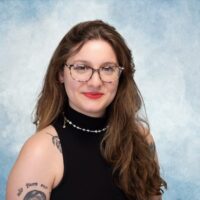 Beatriz é uma mulher branca, de cabelos longos castanhos avermelhados, que usa óculos e tem tatuagens