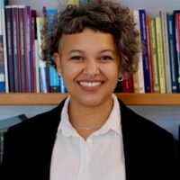 Fernanda é uma mulher negra de pele clara e cabelos castanhos curtos e cacheados. Ela usa uma caminha branca e um blazer preto.