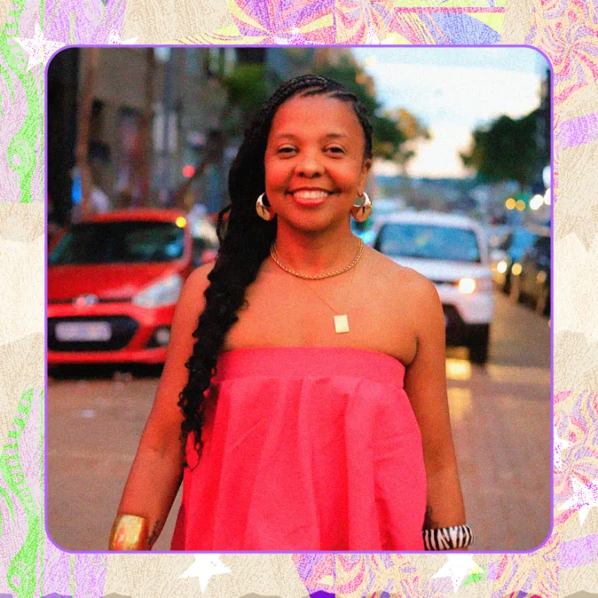 Andreza Ferreira, mulher negra sorridente com vestido rosa, acessórios dourados e trança lateral, posa em rua ao entardecer com carros e luzes ao fundo.