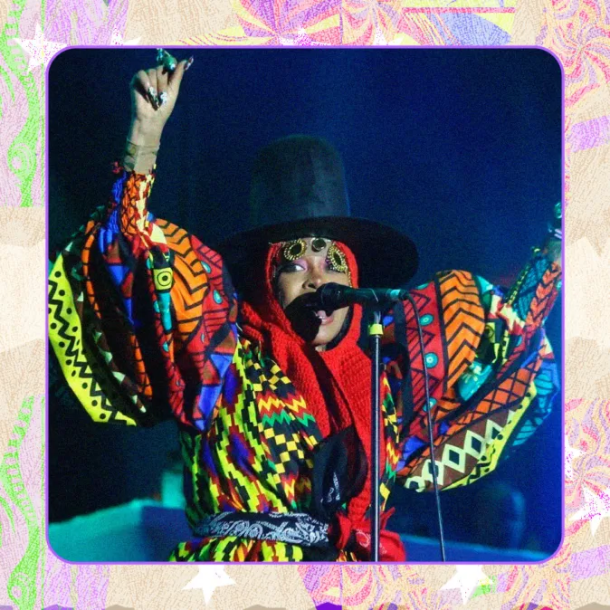 Cantora Erika Baduh no palco com chapéu grande e roupa colorida estampada, braços erguidos e expressão intensa, sobre fundo escuro e moldura colorida.