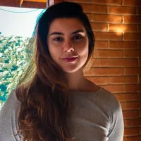 Marina é uma mulher branca de cabelos castanhos longos, usando uma blusa cinza de manga comprida