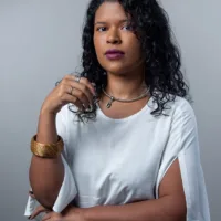 Juliana é uma mulher negra de cabelos cacheados e longos. Ela usa uma blusa branca, colar e bracelete.
