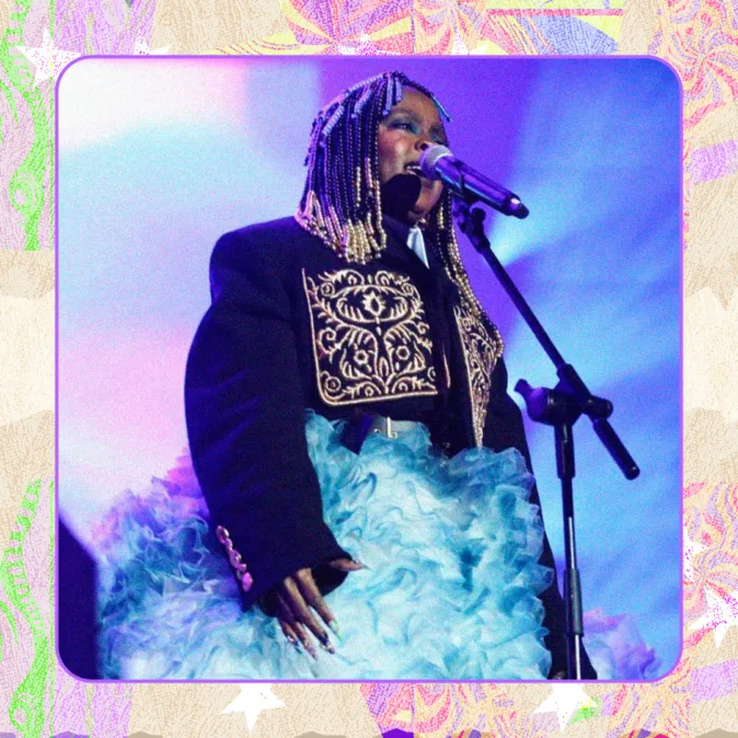 Cantora Lauryn Hill no palco com microfone, usando casaco bordado preto e saia volumosa azul, tranças com miçangas e luz azulada ao fundo.