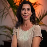 Melina é uma mulher branca de cabelos abaixo dos ombros, vestindo uma roupa bege