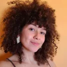 Raquel tem a pele clara, cabelos crespos castanhos avermelhados na altura do ombro e usa brincos grandes.
