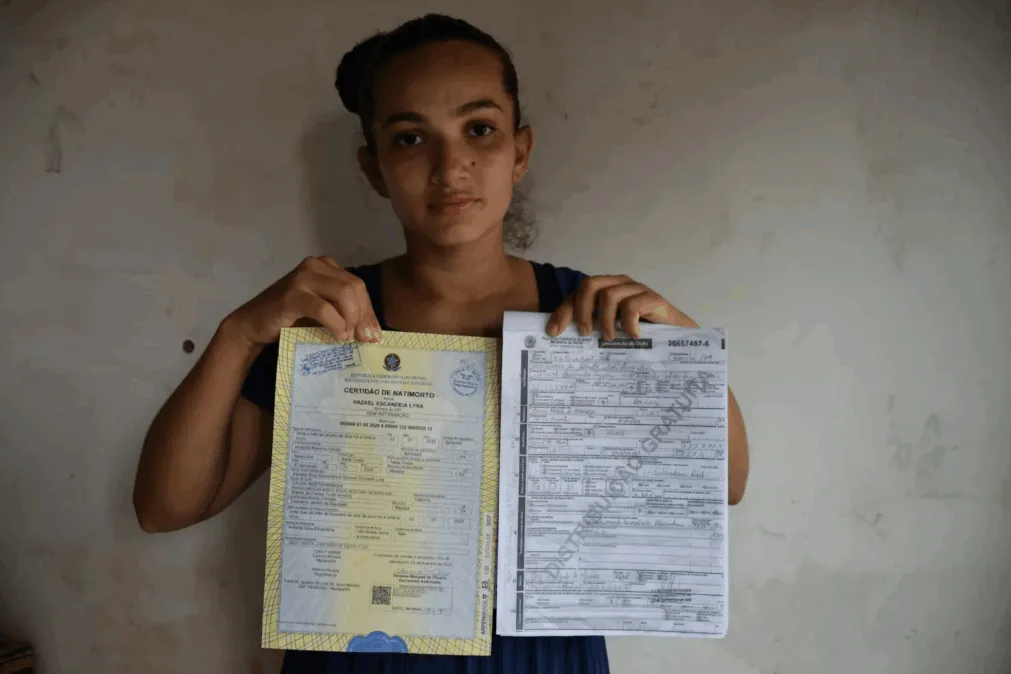Jovem de expressão séria segura certidão de natimorto e declaração de óbito frente a uma parede clara, vestida com blusa azul escura.