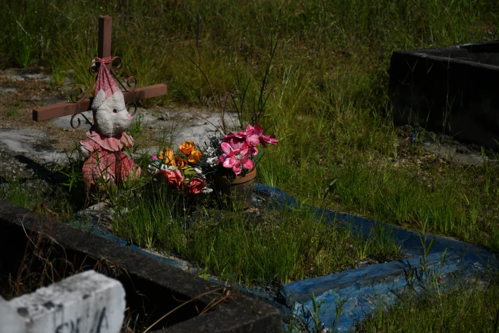 Túmulo infantil com moldura azul e cruz de madeira. Um ursinho de pelúcia vestido marca a lápide, cercado por flores artificiais coloridas.