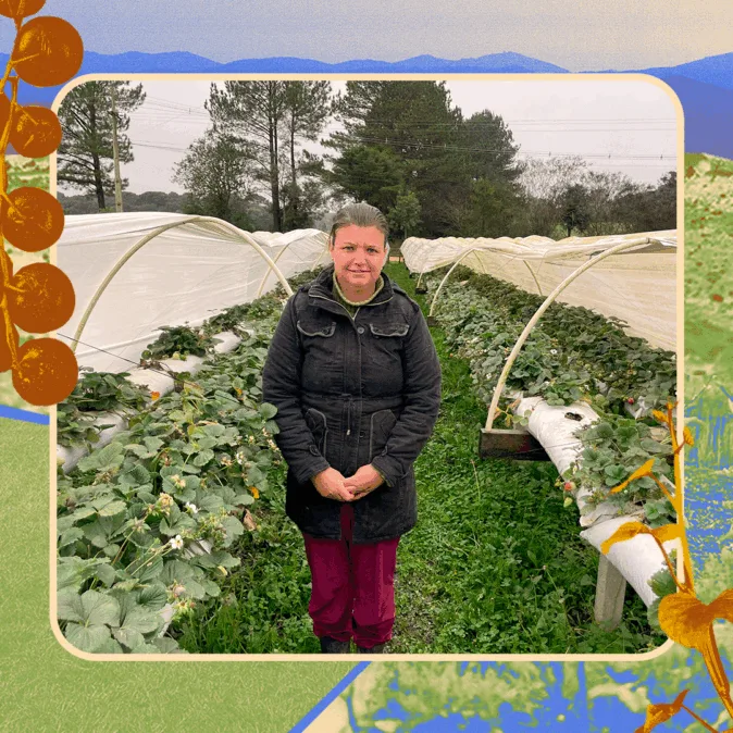 Mulher em estufa de cultivo de morangos, entre fileiras cobertas por lonas plásticas. Ela veste casaco preto e calça bordô, com expressão serena, cercada por vegetação verde e árvores ao fundo. O clima é nublado e frio, com visual rural ao redor.