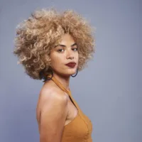 Talita Azevedo é uma mulher negra de pele clara, cabelos crespos loiro, com tamanho médio, ela usa batom escuro e vestido ocre