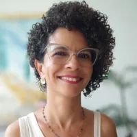Ana Fernanda é uma mulher parda de cabelos cacheados curtos, usa óculos e uma camiseta branca