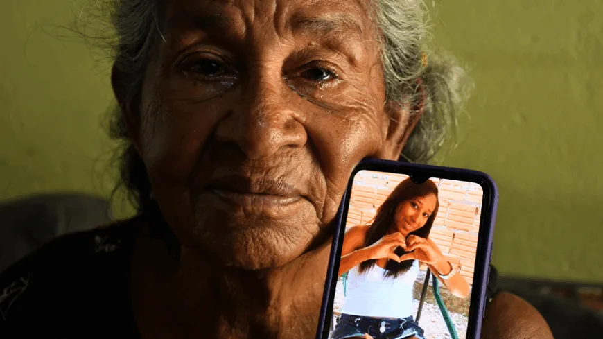 A imagem mostra uma senhora idosa segurando um celular. No celular, aparece a foto de uma jovem fazendo um gesto de coração com as mãos. A senhora tem uma expressão séria e aparenta estar emocionada, com olhos marejados.