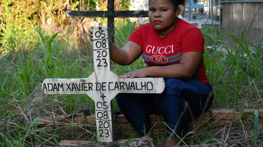 Mulher jovem sentada ao lado de uma cruz branca com o nome “Adam Xavier de Carvalho” em cemitério, expressão séria, vegetação alta ao redor.