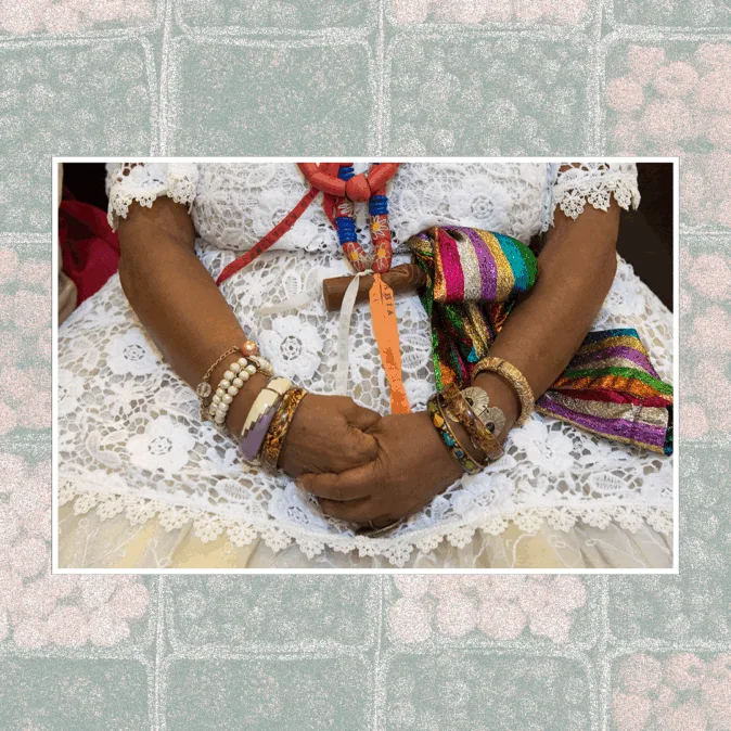 A imagem mostra as mãos de uma mulher negra, cruzadas sobre o colo, vestindo uma roupa branca tradicional de baianas de acarajé. Ela usa várias pulseiras coloridas e fitas de cetim, além de colares de contas e um pano multicolorido. A fotografia está em uma moldura preenchida por padrões de frutas estilizadas.