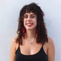 Marina tem pele clara, cabelos crespos castanhos abaixo dos ombros, está sorrindo e usa brincos vermelhos.