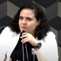Mulher parda clara de cabelos escuros cacheados na altura dos ombros, segurando um microfone.