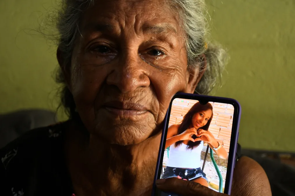 A imagem mostra uma idosa segurando um celular. No celular, aparece a foto de uma jovem fazendo um gesto de coração com as mãos. A senhora tem uma expressão séria e aparenta estar emocionada, com olhos marejados.