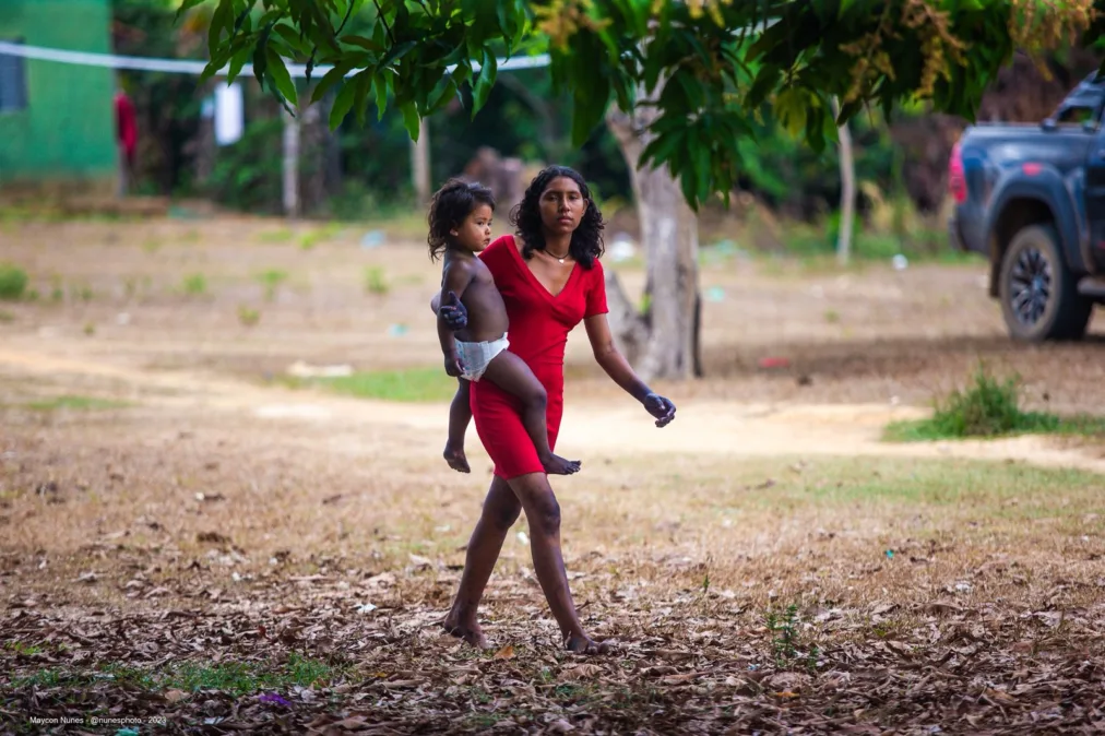 Yapê, uma adolescente indígena de vestido vermelho caminha descalça por um terreno seco, carregando uma criança de fralda no colo.