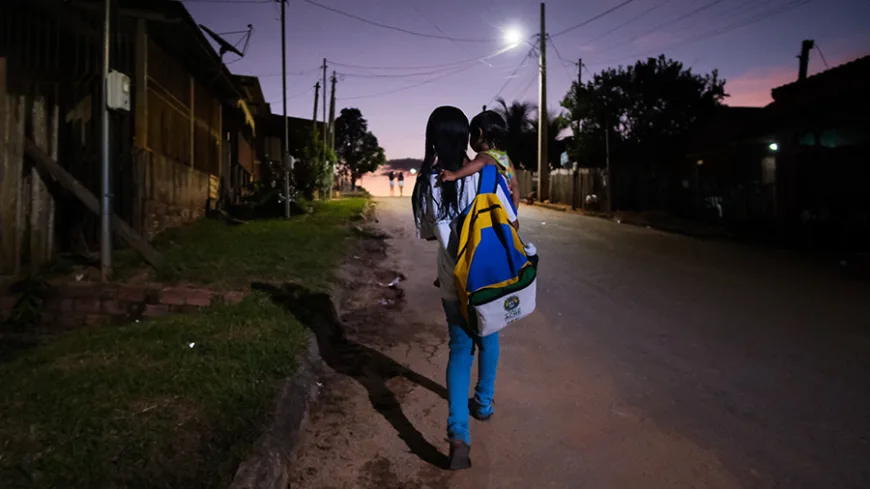 menina caminha com filho no colo e mochila grande da escola nas costas, pelas ruas do municipio do interior ao por do sol