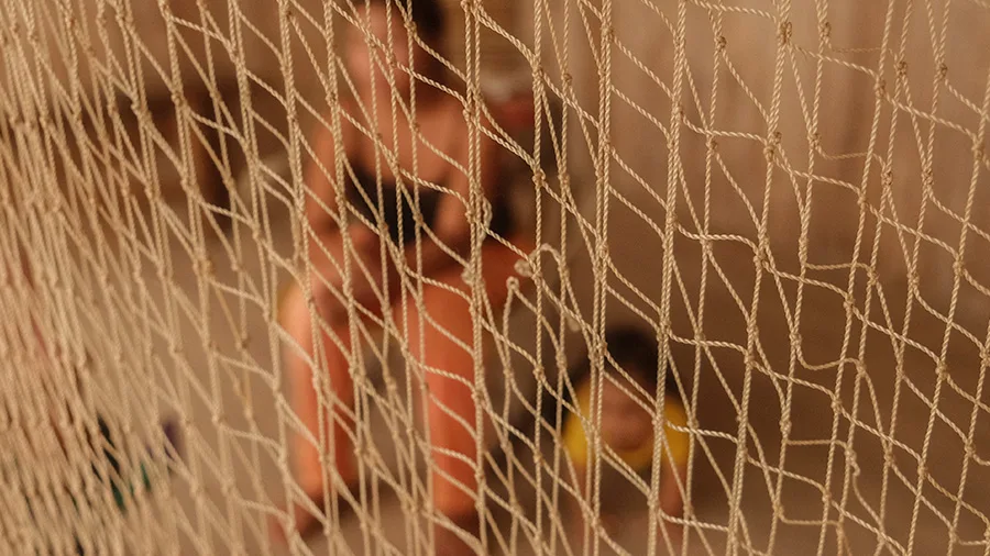 Mulher sentada em uma cadeira ao fundo, desfocada, é vista através de uma rede esticada em primeiro plano. A rede, em foco, ocupa quase toda a imagem, criando uma sensação de separação ou barreira.