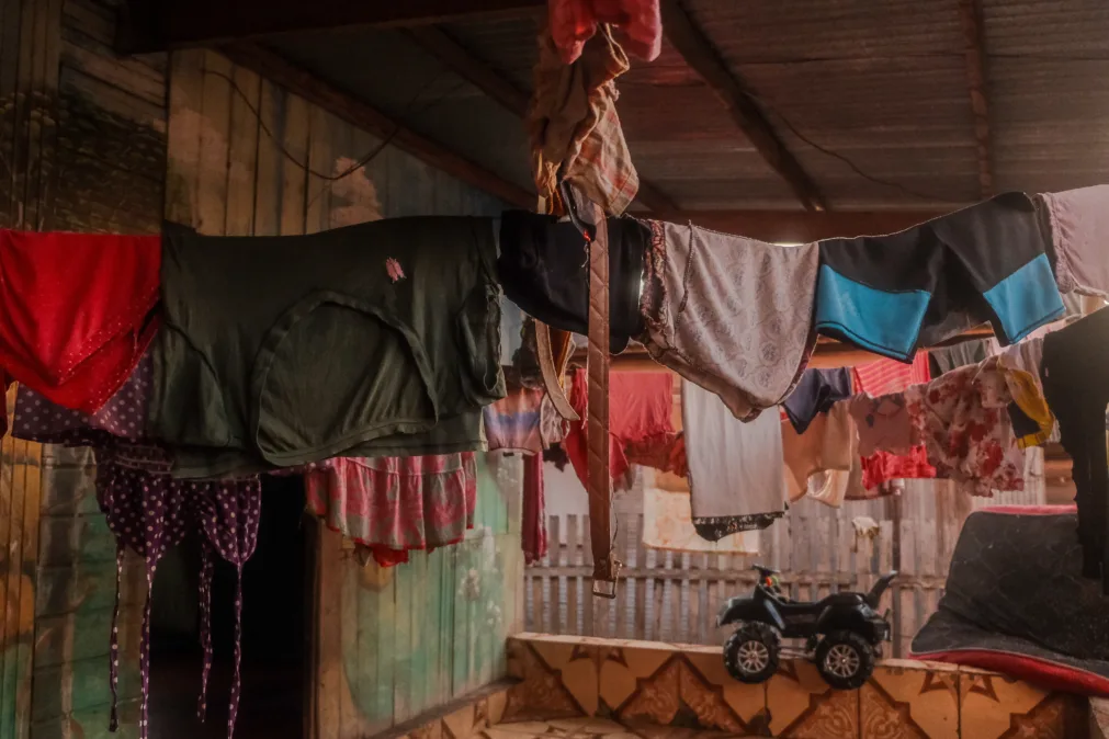 Imagem mostra dezenas de roupas estendidas em varais improvisados dentro de uma casa de madeira. As peças, coloridas e variadas, incluem roupas íntimas penduradas em cordas presas ao teto. Ao fundo, vê-se uma pequena moto de brinquedo e uma cerca de madeira.