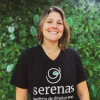 Amanda é uma mulher branca de cabelos loiros na altura dos ombros, ela usa uma blusa preta escrito Serenas e sorri.