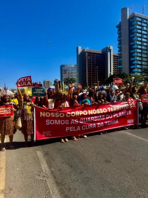 Mulheres indígenas marcham com faixa vermelha que diz: “Nosso corpo, nosso território. Somos as guardiãs do planeta pela cura da terra.”