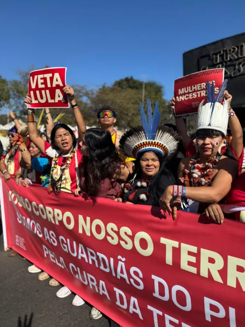 Mulheres indígenas marcham segurando faixa “Nosso corpo, nosso território” e cartazes “Veta Lula” e “Mulheres Ancestrais” em ato de protesto.