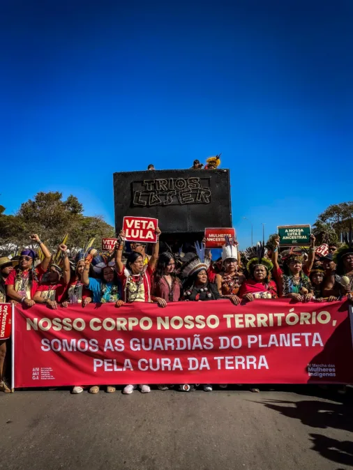 Mulheres indígenas marcham com faixa vermelha que diz: “Nosso corpo, nosso território. Somos as guardiãs do planeta pela cura da terra.”