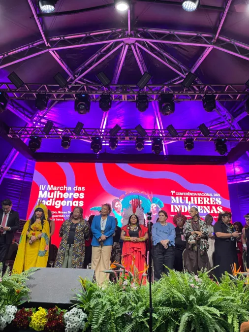 Grupo de mulheres indígenas e apoiadores no palco da IV Marcha e 1ª Conferência Nacional das Mulheres Indígenas, diante de telão vermelho e roxo.