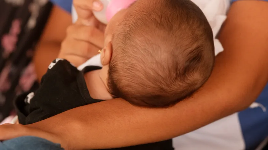Menina dá mamadeira para bebê nos braços dela sentada. Ela veste camiseta com estampa de símbolo institucional e calça jeans.