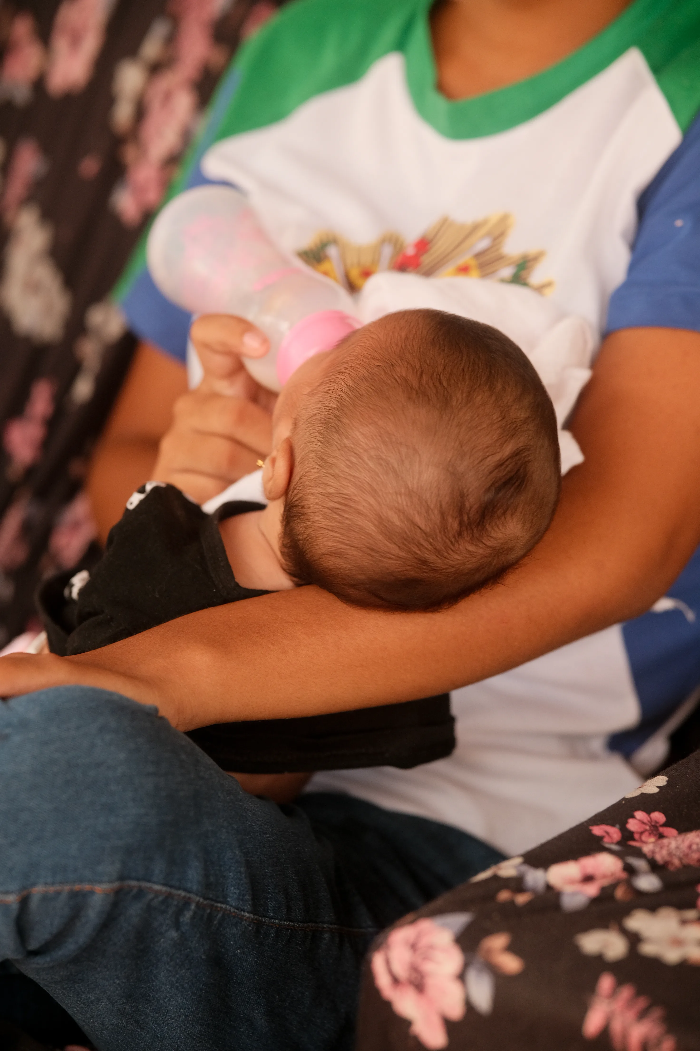 Menina dá mamadeira para bebê nos braços dela sentada. Ela veste camiseta com estampa de símbolo institucional e calça jeans.