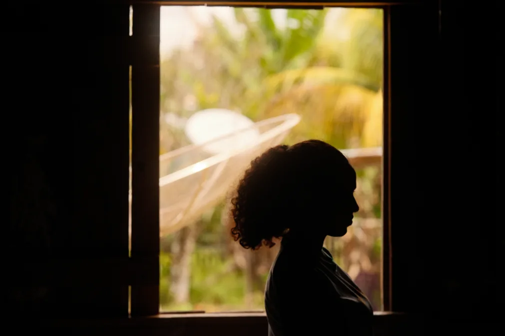 Silhueta de uma menina em perfil aparece contra a luz natural que entra por uma janela. Ao fundo, vê-se vegetação tropical desfocada, com tons verdes e amarelos.