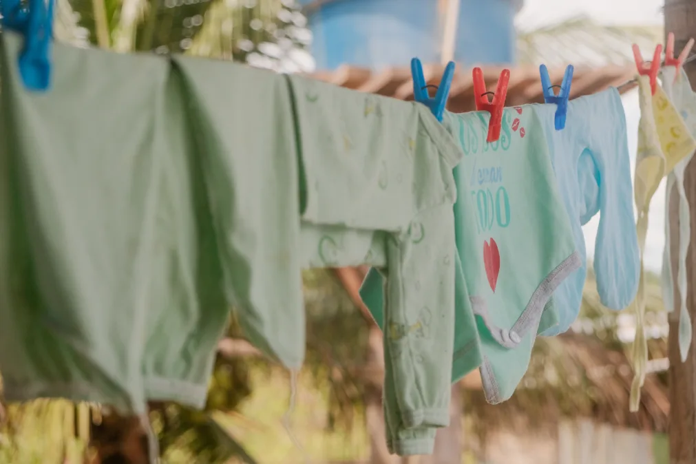 Roupas de bebê penduradas em um varal com pregadores coloridos, secam ao ar livre. As peças, em tons suaves de verde, azul e amarelo, trazem delicadeza à cena, com destaque para um body estampado com um coração vermelho. 