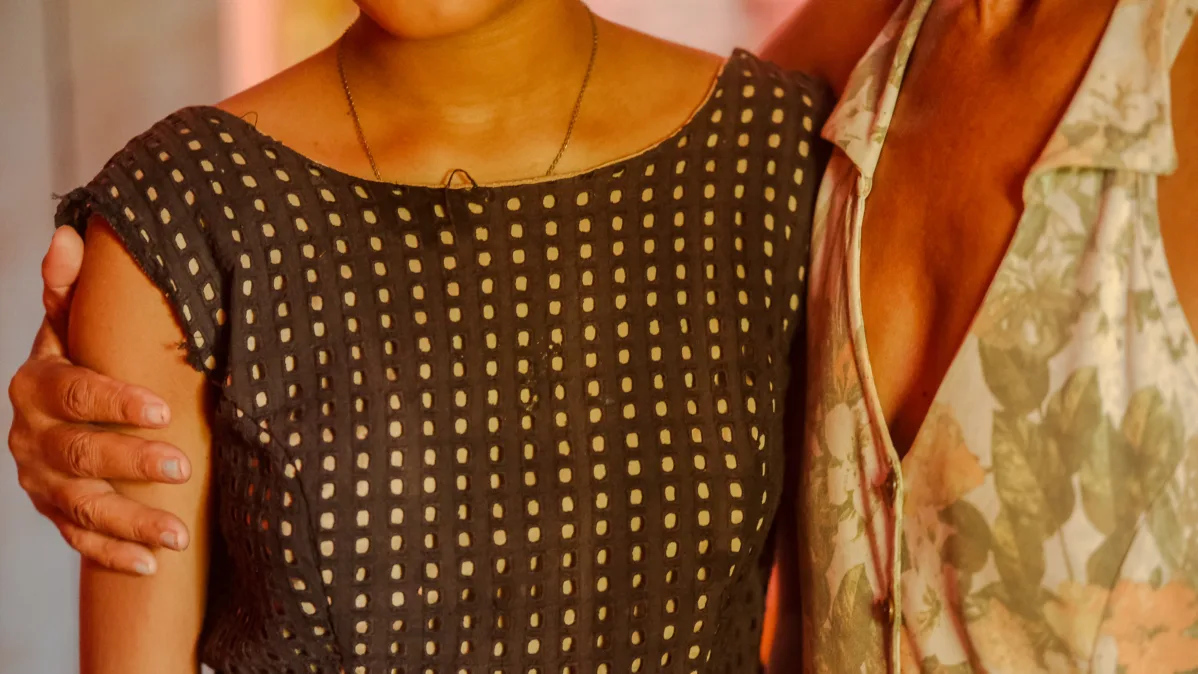 Duas mulheres se abraçam lado a lado. O enquadramento destaca seus troncos e roupas: uma veste blusa escura com padrão de pequenos quadrados claros; a outra, vestido floral claro com decote transpassado. 