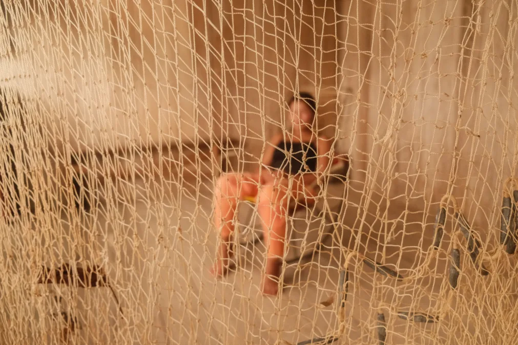 Mulher sentada em uma cadeira ao fundo, desfocada, é vista através de uma rede esticada em primeiro plano. A rede, em foco, ocupa quase toda a imagem, criando uma sensação de separação ou barreira.