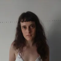 Raabe é uma mulher branca de cabelos ruivos longos e com franja, ela usa uma camiseta branca