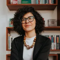 Maria é uma mulher de pele clara, cabelos cacheados acima dos ombros, que usa óculos, um colar de madeira, blusa branca e blazer preto