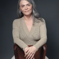 Netta é uma mulher branca de cabelos grisalhos longos, ela tem uma expressão serena e usa uma blusa bege