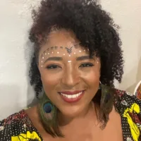 Cecília é uma mulher negra de cabelos crespos de tamanho médio, ela usa uma pintura acima dos olhos e sorri
