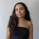 Amanda é uma mulher parda de cabelos castanhos longos, ela usa uma blusa tomara-que-caia preta e tem uma expressão leve
